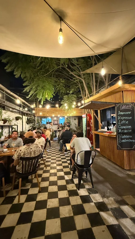 Vista del patio lleno de noche, con el piso ajedrez y la pizarra de la cafetería al fondo.